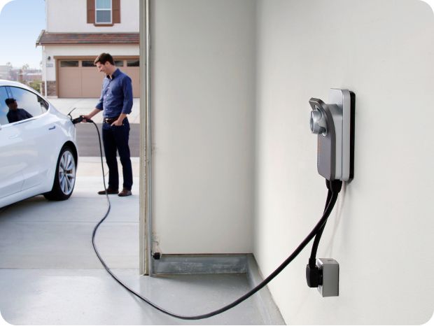 Mies lataamassa valkoista sähköautoa kotona autotallin seinässä olevalla latausasemalla.